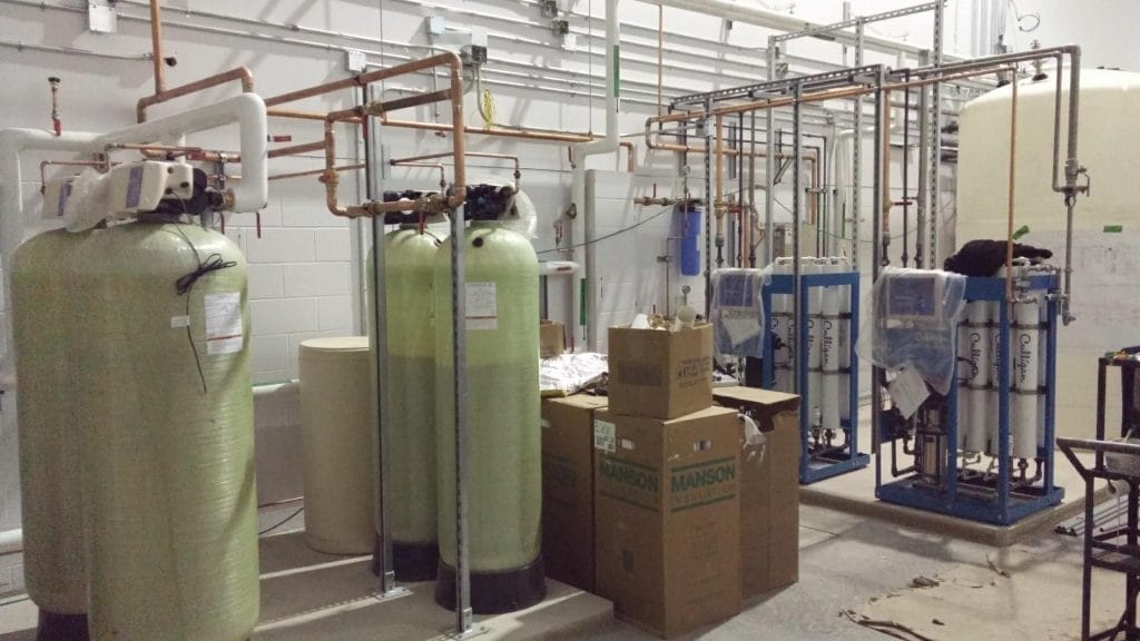 "Système industriel de traitement de l'eau Culligan installé dans une salle technique, avec de grands réservoirs verts, des filtres, et des tuyaux en cuivre. Des cartons de fournitures sont également visibles."